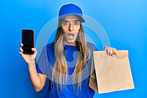 Young hispanic woman holding delivery paper bag and showing smartphone screen afraid and shocked with surprise and amazed