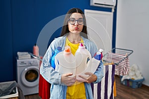 Young hispanic woman holding detergent bottles puffing cheeks with funny face