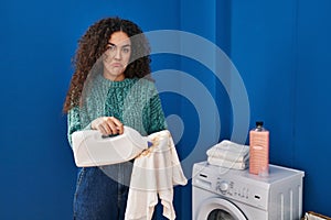 Young hispanic woman holding dirty laundry and detergent bottle depressed and worry for distress, crying angry and afraid