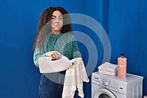 Young hispanic woman holding dirty laundry and detergent bottle puffing cheeks with funny face