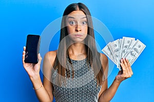 Young hispanic woman holding dollars and using smarpthone puffing cheeks with funny face