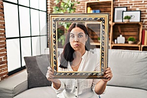 Young hispanic woman holding empty frame puffing cheeks with funny face