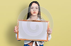Young hispanic woman holding empty white chalkboard puffing cheeks with funny face