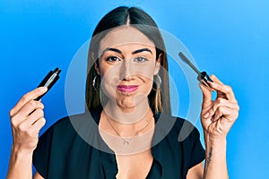 Young hispanic woman holding eyelash mascara puffing cheeks with funny face