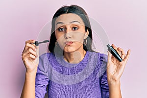 Young hispanic woman holding eyelashes curler puffing cheeks with funny face