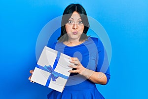 Young hispanic woman holding gift puffing cheeks with funny face