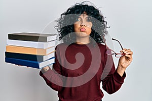 Young hispanic woman holding glasses and books puffing cheeks with funny face