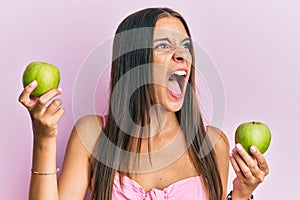 Young hispanic woman holding green apples angry and mad screaming frustrated and furious, shouting with anger