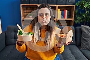 Young hispanic woman holding healthy salad and fried chicken wings puffing cheeks with funny face