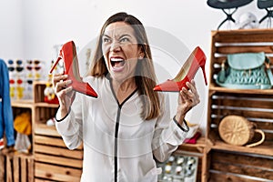 Young hispanic woman holding high heel shoes at clothing store angry and mad screaming frustrated and furious, shouting with anger