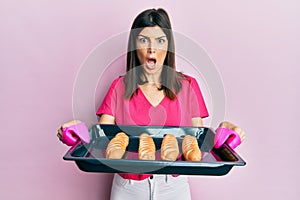 Young hispanic woman holding homemade bread afraid and shocked with surprise and amazed expression, fear and excited face