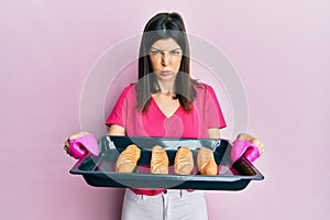 Young hispanic woman holding homemade bread puffing cheeks with funny face