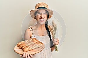 Young hispanic woman holding homemade bread and spike wheat sticking tongue out happy with funny expression