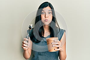Young hispanic woman holding ice cream puffing cheeks with funny face