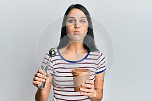 Young hispanic woman holding ice cream puffing cheeks with funny face