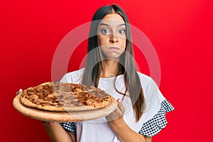 Young hispanic woman holding italian pizza puffing cheeks with funny face