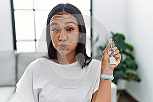 Young hispanic woman holding keys of new home puffing cheeks with funny face