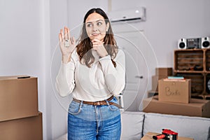 Young hispanic woman holding keys of new home serious face thinking about question with hand on chin, thoughtful about confusing