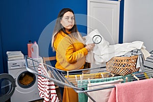 Young hispanic woman holding magnifying glass looking for stain at clothes puffing cheeks with funny face