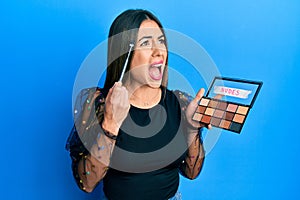 Young hispanic woman holding makeup brush and blush angry and mad screaming frustrated and furious, shouting with anger