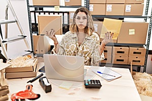 Young hispanic woman holding package and envelope sitting on desk puffing cheeks with funny face