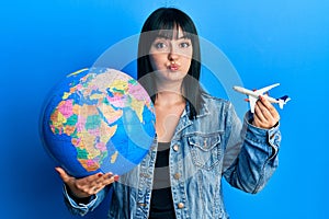 Young hispanic woman holding paper toy and world ball puffing cheeks with funny face