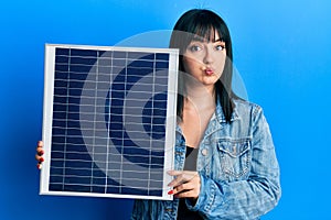 Young hispanic woman holding photovoltaic solar panel puffing cheeks with funny face