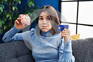 Young hispanic woman holding piggy bank and credit card puffing cheeks with funny face