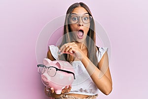 Young hispanic woman holding piggy bank with glasses and coin afraid and shocked with surprise and amazed expression, fear and