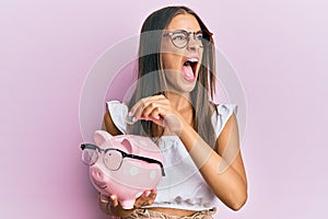 Young hispanic woman holding piggy bank with glasses and coin angry and mad screaming frustrated and furious, shouting with anger