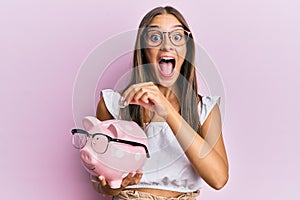 Young hispanic woman holding piggy bank with glasses and coin celebrating crazy and amazed for success with open eyes screaming