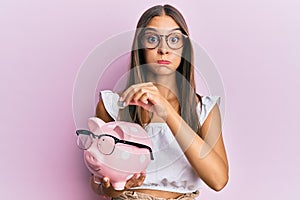 Young hispanic woman holding piggy bank with glasses and coin puffing cheeks with funny face