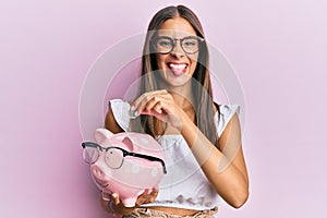 Young hispanic woman holding piggy bank with glasses and coin sticking tongue out happy with funny expression