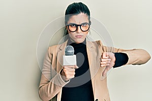 Young hispanic woman holding reporter microphone with angry face, negative sign showing dislike with thumbs down, rejection