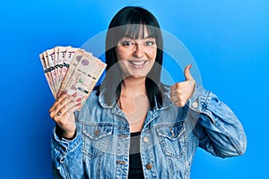 Young hispanic woman holding saudi arabia riyal banknotes smiling happy and positive, thumb up doing excellent and approval sign