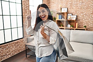 Young hispanic woman holding shopping bags smiling with an idea or question pointing finger with happy face, number one