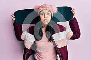 Young hispanic woman holding skate puffing cheeks with funny face