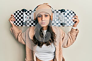 Young hispanic woman holding skate wearing headphones puffing cheeks with funny face