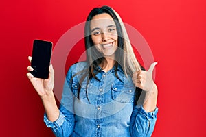 Young hispanic woman holding smartphone showing screen smiling happy and positive, thumb up doing excellent and approval sign