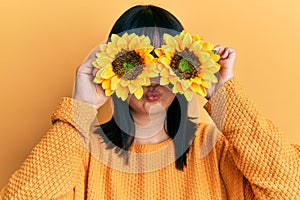 Young hispanic woman holding sunflowers over eyes puffing cheeks with funny face