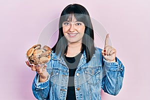 Young hispanic woman holding sweet pastries smiling with an idea or question pointing finger with happy face, number one