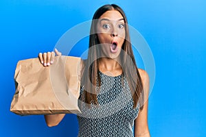 Young hispanic woman holding take away paper bag scared and amazed with open mouth for surprise, disbelief face