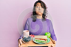 Young hispanic woman holding tray with breakfast food puffing cheeks with funny face