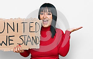 Young hispanic woman holding united we stand banner celebrating victory with happy smile and winner expression with raised hands