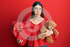 Young hispanic woman holding valentine gift puffing cheeks with funny face