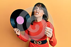 Young hispanic woman holding vinyl disc annoyed and frustrated shouting with anger, yelling crazy with anger and hand raised