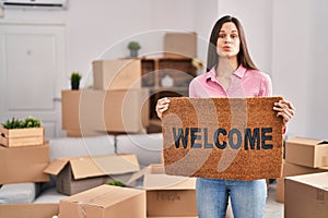 Young hispanic woman holding welcome doormat at new home puffing cheeks with funny face