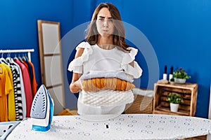 Young hispanic woman ironing clothes at laundry room holding folded sweaters puffing cheeks with funny face