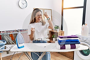 Young hispanic woman ironing clothes at laundry room very happy and excited doing winner gesture with arms raised, smiling and