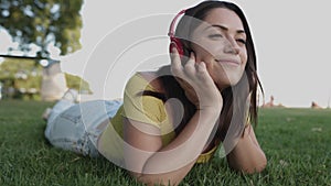 Young hispanic woman listening to music with headphones while relaxing on green grass at park